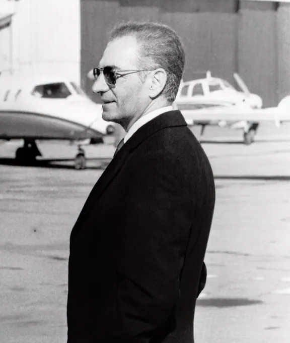 His Majesty, the Shah of Iran in a dark suit and sunglasses stands in profile on an airfield with planes — a public domain photograph from The Shah of Iran archive documenting the Pahlavi era.