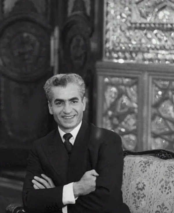His Majesty, the Shah of Iran in a dark suit and tie sits with crossed arms against an ornate background — a public domain photograph from The Shah of Iran archive documenting the Pahlavi era.