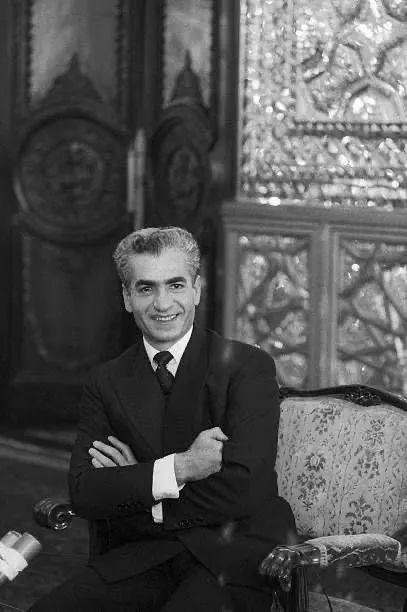 His Majesty, the Shah of Iran in a dark suit and tie sits with arms crossed in an ornate room — a public domain photograph from The Shah of Iran archive documenting the Pahlavi era.