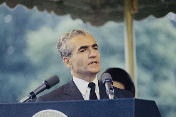 His Majesty, the Shah of Iran in a dark suit and tie speaks into two microphones at a podium — a public domain photograph from The Shah of Iran archive documenting the Pahlavi era.