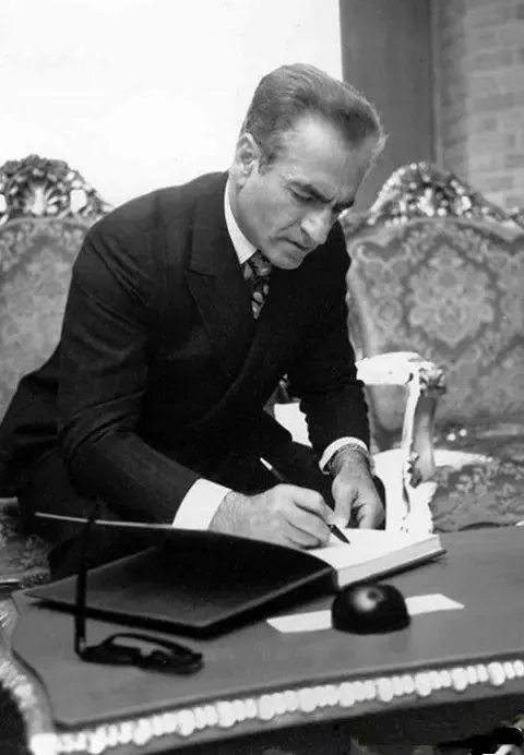 His Majesty, the Shah of Iran in a dark suit sits at a desk and writes in a book — a public domain photograph from The Shah of Iran archive documenting the Pahlavi era.