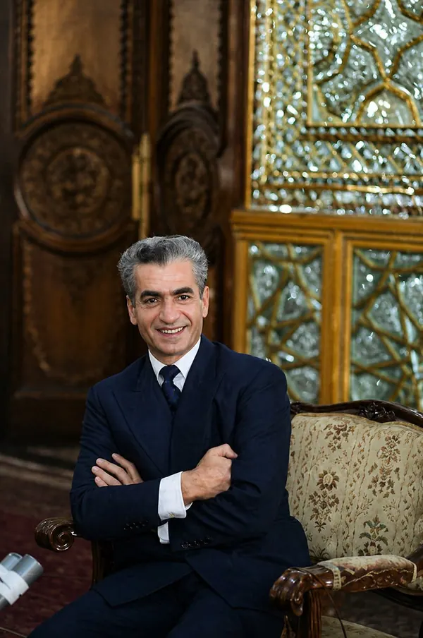 His Majesty, the Shah of Iran in a dark suit sits with crossed arms in an ornate room — a public domain photograph from The Shah of Iran archive documenting the Pahlavi era.