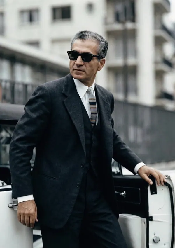 His Majesty, the Shah of Iran in a dark three-piece suit and sunglasses leans against a white car outdoors — a public domain photograph from The Shah of Iran archive documenting the Pahlavi era.