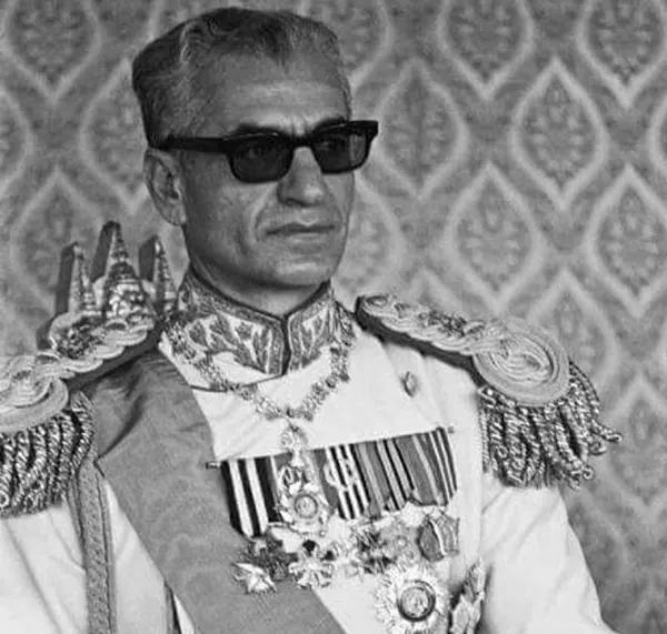 His Majesty, the Shah of Iran in a decorated military uniform and dark sunglasses poses against a patterned wall — a public domain photograph from The Shah of Iran archive documenting the Pahlavi era.