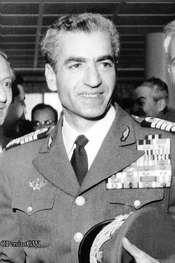 His Majesty, the Shah of Iran in a decorated military uniform smiles while holding a peaked cap in his hand — a public domain photograph from The Shah of Iran archive documenting the Pahlavi era.