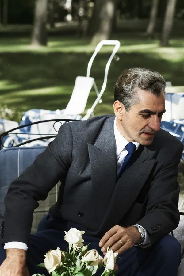 His Majesty, the Shah of Iran in a grey suit and blue tie sits outdoors near white flowers — a public domain photograph from The Shah of Iran archive documenting the Pahlavi era.