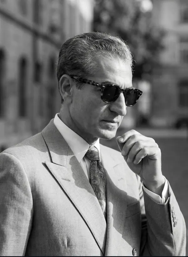 His Majesty, the Shah of Iran in a light-colored suit and sunglasses looks downward with his hand near his chin — a public domain photograph from The Shah of Iran archive documenting the Pahlavi era.