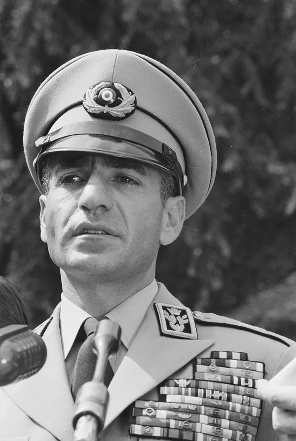 His Majesty, the Shah of Iran in a military uniform and peaked cap speaks into microphones against a blurred background — a public domain photograph from The Shah of Iran archive documenting the Pahlavi era.