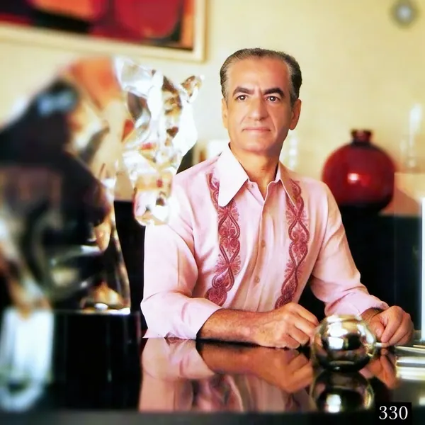 His Majesty, the Shah of Iran in a pink embroidered shirt stands behind a reflective surface next to a glass sculpture — a public domain photograph from The Shah of Iran archive documenting the Pahlavi era.