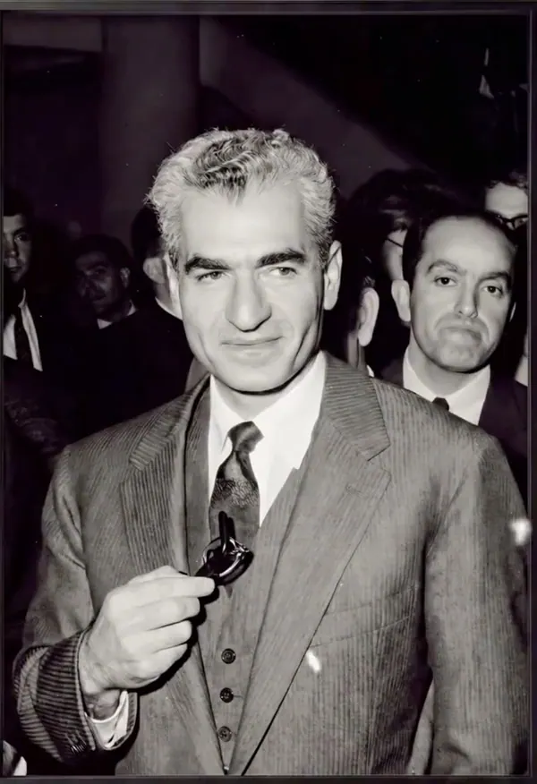 His Majesty, the Shah of Iran in a pinstripe suit and tie holds a small object in his hand — a public domain photograph from The Shah of Iran archive documenting the Pahlavi era.