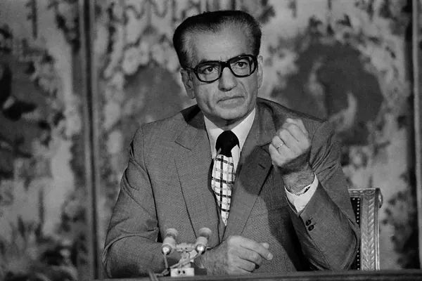 His Majesty, the Shah of Iran in a suit and glasses gestures with his hand while sitting behind microphones — a public domain photograph from The Shah of Iran archive documenting the Pahlavi era.