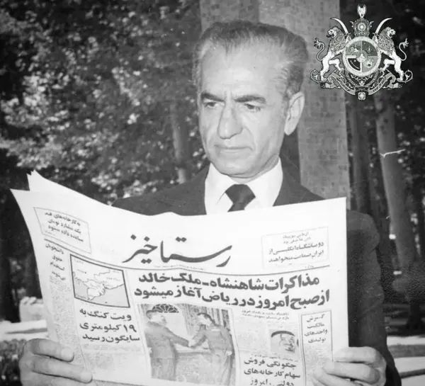 His Majesty, the Shah of Iran in a suit and tie holds and reads a large newspaper outdoors — a public domain photograph from The Shah of Iran archive documenting the Pahlavi era.