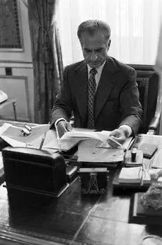 His Majesty, the Shah of Iran in a suit and tie sits at a desk reviewing several papers — a public domain photograph from The Shah of Iran archive documenting the Pahlavi era.