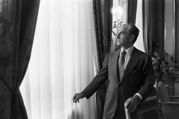 His Majesty, the Shah of Iran in a suit and tie stands by a window, looking out through curtains — a public domain photograph from The Shah of Iran archive documenting the Pahlavi era.