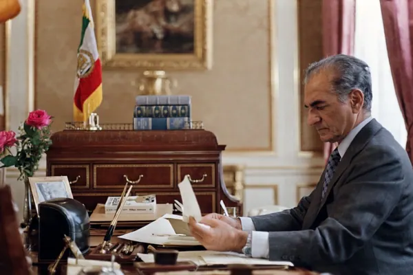His Majesty, the Shah of Iran in a suit sits at a desk reading a piece of paper in an office — a public domain photograph from The Shah of Iran archive documenting the Pahlavi era.