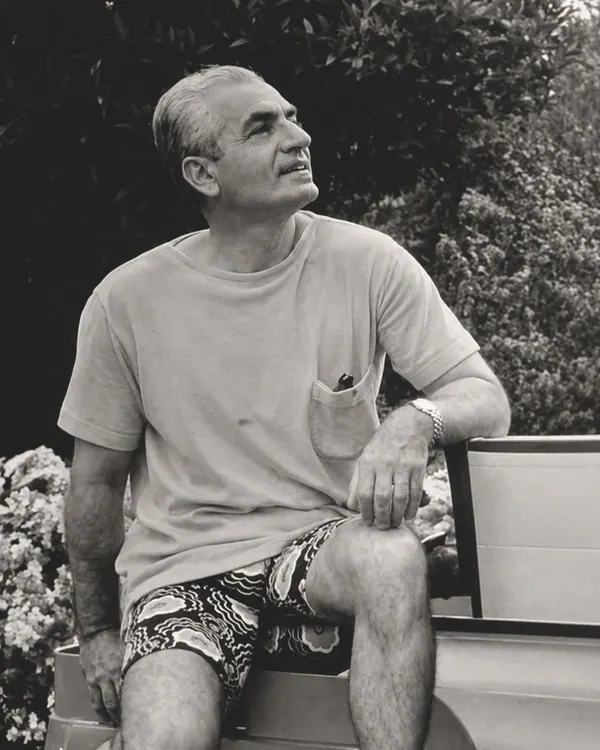 His Majesty, the Shah of Iran in a t-shirt and patterned shorts sits outdoors against a backdrop of foliage — a public domain photograph from The Shah of Iran archive documenting the Pahlavi era.