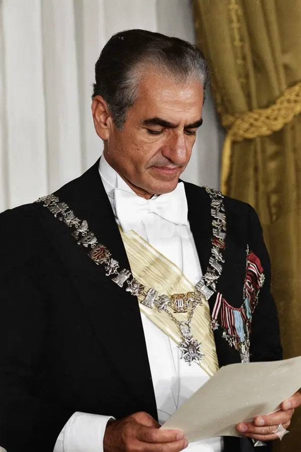 His Majesty, the Shah of Iran in a tuxedo and ceremonial collar reads a piece of paper — a public domain photograph from The Shah of Iran archive documenting the Pahlavi era.