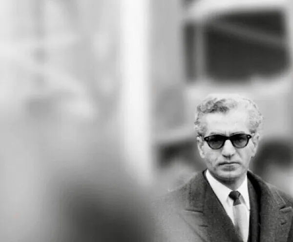 His Majesty, the Shah of Iran wearing dark sunglasses, a suit, and a tie looks forward against a blurred background — a public domain photograph from The Shah of Iran archive documenting the Pahlavi era.