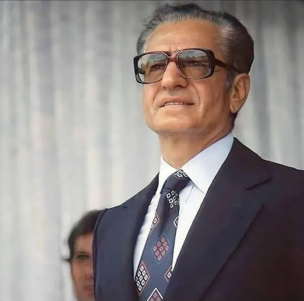 His Majesty, the Shah of Iran wearing glasses, a dark suit, and a patterned tie looks upward against a curtained background — a public domain photograph from The Shah of Iran archive documenting the Pahlavi era.