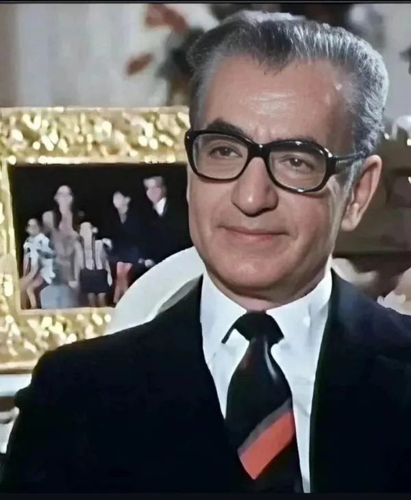 His Majesty, the Shah of Iran wearing glasses, a dark suit, and a tie poses before a gold-framed picture — a public domain photograph from The Shah of Iran archive documenting the Pahlavi era.