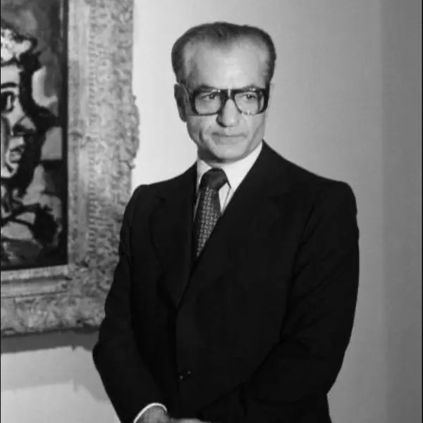 His Majesty, the Shah of Iran wearing glasses and a dark suit stands beside a framed painting — a public domain photograph from The Shah of Iran archive documenting the Pahlavi era.