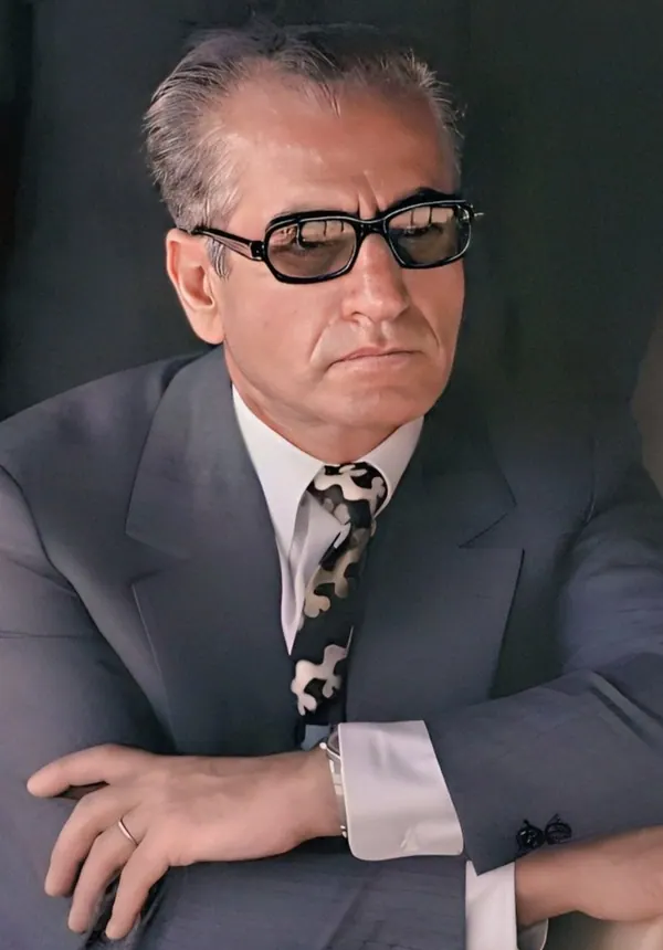 His Majesty, the Shah of Iran wearing sunglasses, a suit, and a patterned tie poses with arms crossed — a public domain photograph from The Shah of Iran archive documenting the Pahlavi era.