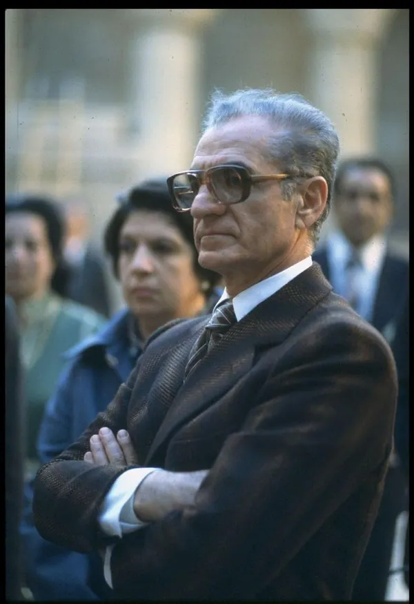 An older man in a brown suit and glasses stands with arms crossed among other people — a public domain photograph from The Shah of Iran archive documenting the Pahlavi era.