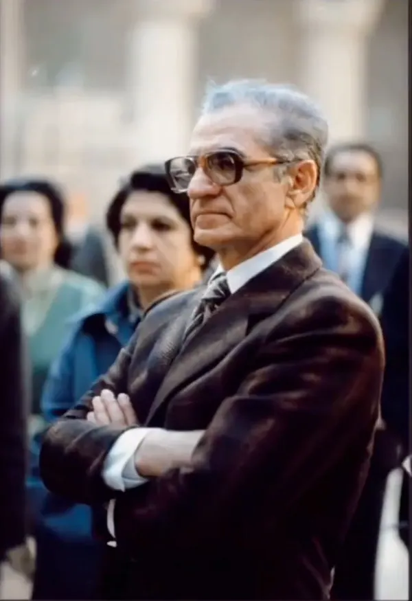 An older man in a dark suit and glasses stands with arms crossed, flanked by other people — a public domain photograph from The Shah of Iran archive documenting the Pahlavi era.