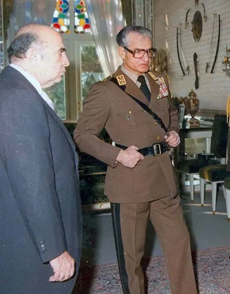 An older man in a dark suit stands facing His Majesty in a brown military uniform with medals — a public domain photograph from The Shah of Iran archive documenting the Pahlavi era.