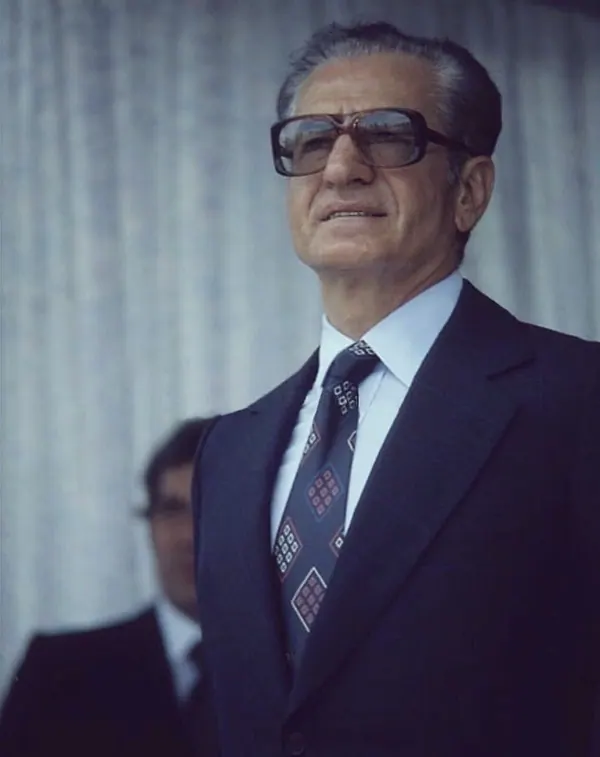 An older man in a dark suit, tie, and sunglasses looks upward against a light curtain background — a public domain photograph from The Shah of Iran archive documenting the Pahlavi era.