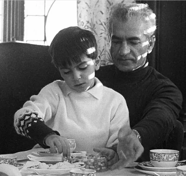 An older man in a dark sweater watches a young boy in a white sweater at a table — a public domain photograph from The Shah of Iran archive documenting the Pahlavi era.