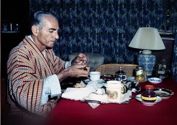 An older man in a patterned robe sits at a red table with tea and food items — a public domain photograph from The Shah of Iran archive documenting the Pahlavi era.