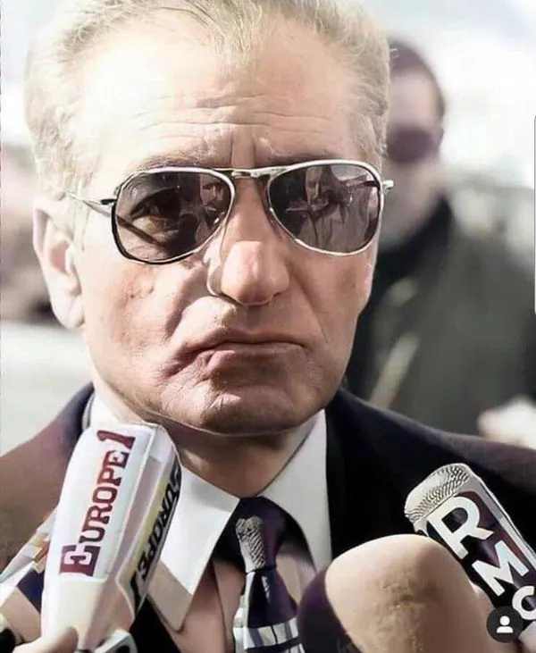 An older man in a suit and sunglasses stands before several microphones during a press event — a public domain photograph from The Shah of Iran archive documenting the Pahlavi era.
