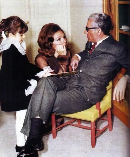 An older man in a suit sits in a chair while Farah Pahlavi, Empress of Iran, and a child look at him — a public domain photograph from The Shah of Iran archive documenting the Pahlavi era.