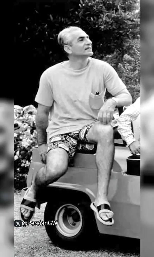 An older man in a t-shirt and patterned shorts sits on the hood of a vehicle — a public domain photograph from The Shah of Iran archive documenting the Pahlavi era.