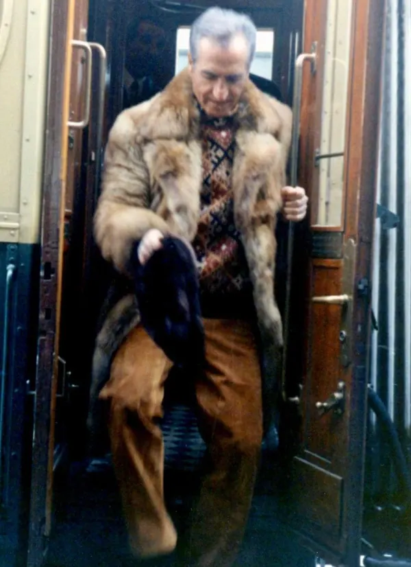 An older man wearing a fur coat and brown trousers steps out of a wooden train carriage — a public domain photograph from The Shah of Iran archive documenting the Pahlavi era.