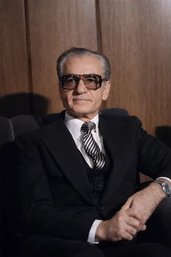 An older man wearing glasses, a dark suit, and a striped tie sits against a wood-paneled wall — a public domain photograph from The Shah of Iran archive documenting the Pahlavi era.