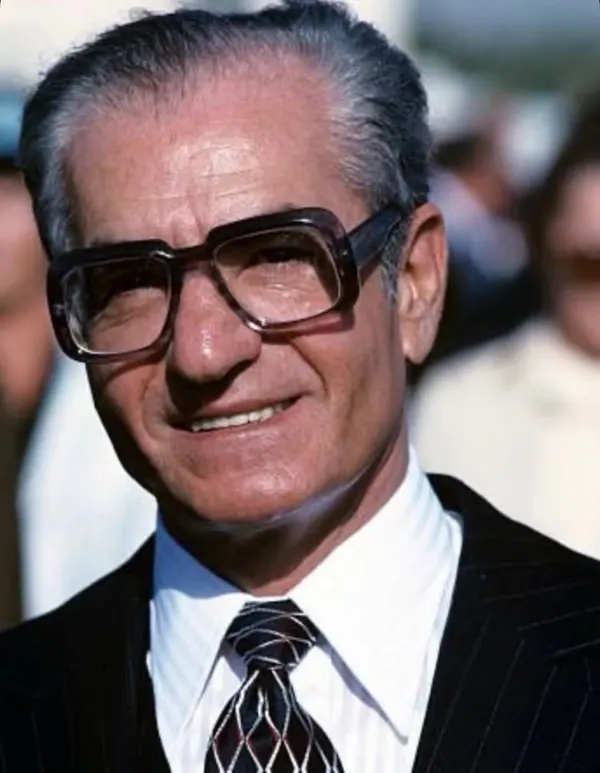 An older man with glasses, a white shirt, and a dark suit smiles for a close-up portrait — a public domain photograph from The Shah of Iran archive documenting the Pahlavi era.