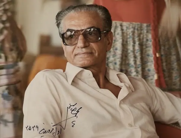 An older man with glasses wearing a light-colored button-down shirt sits in a relaxed posture indoors — a public domain photograph from The Shah of Iran archive documenting the Pahlavi era.