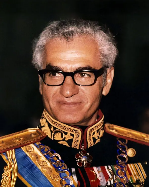 An older man with grey hair and glasses wears an ornate military uniform with gold embroidery and medals — a public domain photograph from The Shah of Iran archive documenting the Pahlavi era.