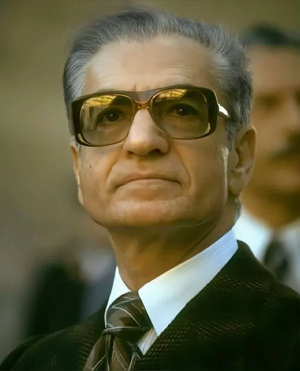 An older man with grey hair and tinted glasses wears a suit and tie in a close-up portrait — a public domain photograph from The Shah of Iran archive documenting the Pahlavi era.