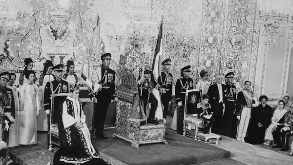 People in formal attire and military uniforms stand around an ornate throne in a decorated room — a public domain photograph from The Shah of Iran archive documenting the Pahlavi era.