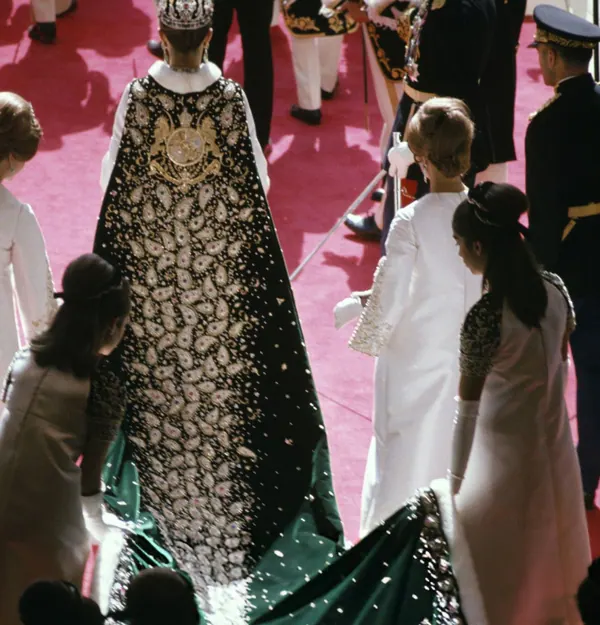People in formal attire and uniforms stand on a red carpet, with one person wearing a long embroidered cape — a public domain photograph from The Shah of Iran archive documenting the Pahlavi era.
