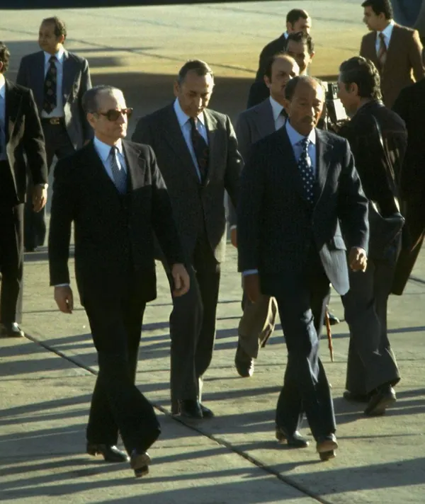Several men in dark business suits walk across a paved outdoor area in bright sunlight — a public domain photograph from The Shah of Iran archive documenting the Pahlavi era.