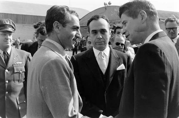 Several men in suits and uniforms stand outdoors, with two men shaking hands in the foreground — a public domain photograph from The Shah of Iran archive documenting the Pahlavi era.