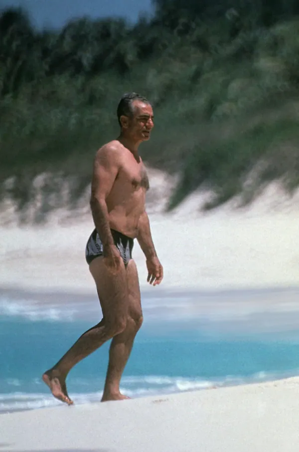 A shirtless middle-aged man in dark swim trunks walks along a white sandy beach by the ocean — a public domain photograph from The Shah of Iran archive documenting the Pahlavi era.