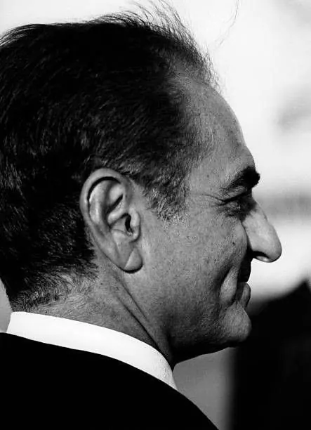 A side profile portrait of His Majesty wearing a dark suit and white collar — a public domain photograph from The Shah of Iran archive documenting the Pahlavi era.