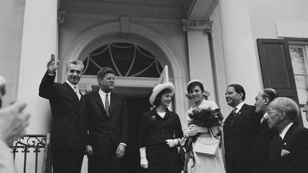 Six people in formal attire stand before an arched doorway, with one man waving his hand — a public domain photograph from The Shah of Iran archive documenting the Pahlavi era.