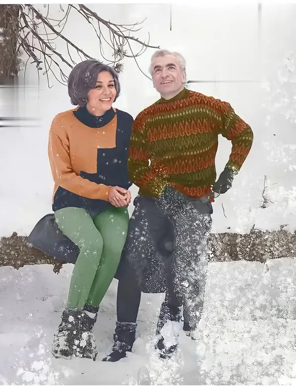 A smiling man and woman in winter clothing sit together outdoors in a snowy landscape — a public domain photograph from The Shah of Iran archive documenting the Pahlavi era.
