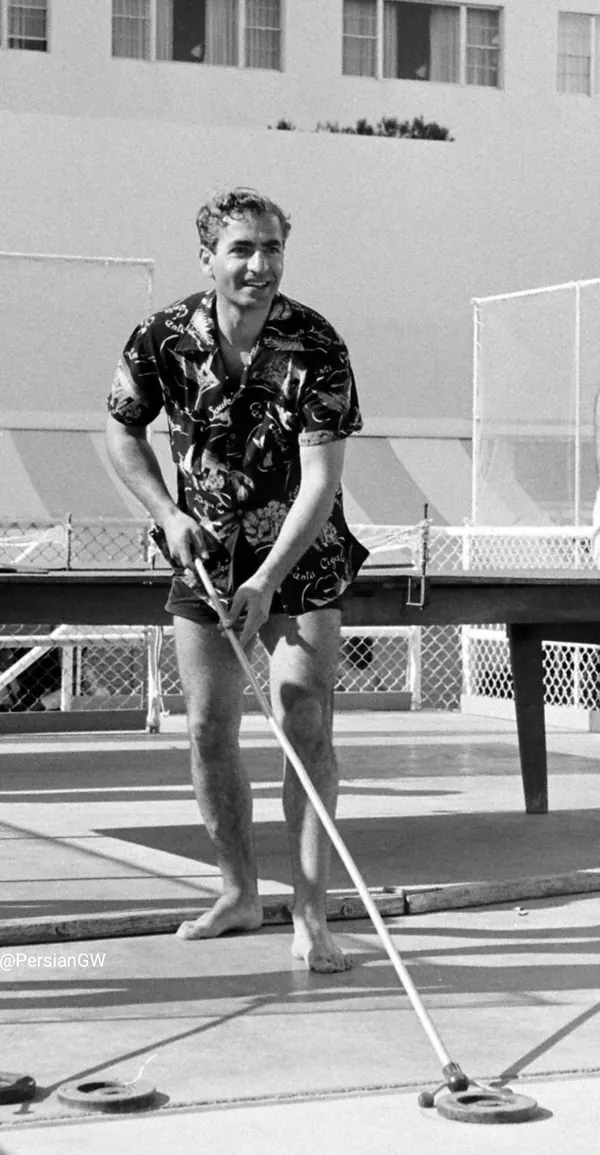 A smiling man in a floral shirt and shorts holds a long pole on a paved outdoor area — a public domain photograph from The Shah of Iran archive documenting the Pahlavi era.